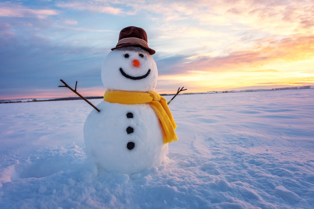A photo of snowman on an icy landscape, with a yellow scarf and a black hat and a large smile with a sunset in the background