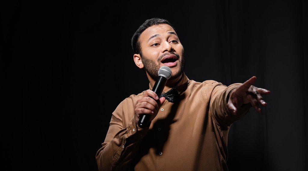 A man with a microphone, pointing to the audience with a black background behind him