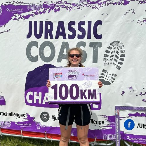 Woman smiling and holding a sign that says '100km', with a backdrop that says 'Jurassic Coast Challenge'