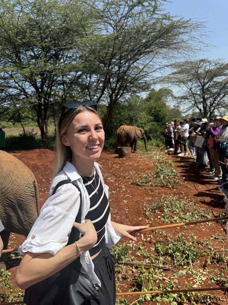 Young white women smiles at camera, with two elephants standing behind her in a sanctuary. The soil is orange and the trees around them are tall and sparse. To the right of the woman are other tourists, taking photos of the elephants.