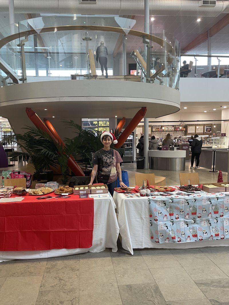 Photo of a young female student, standing behind two stalls laden with baked goods. She wears a Santa hat. Behind her is a cafe and reception area in a university.