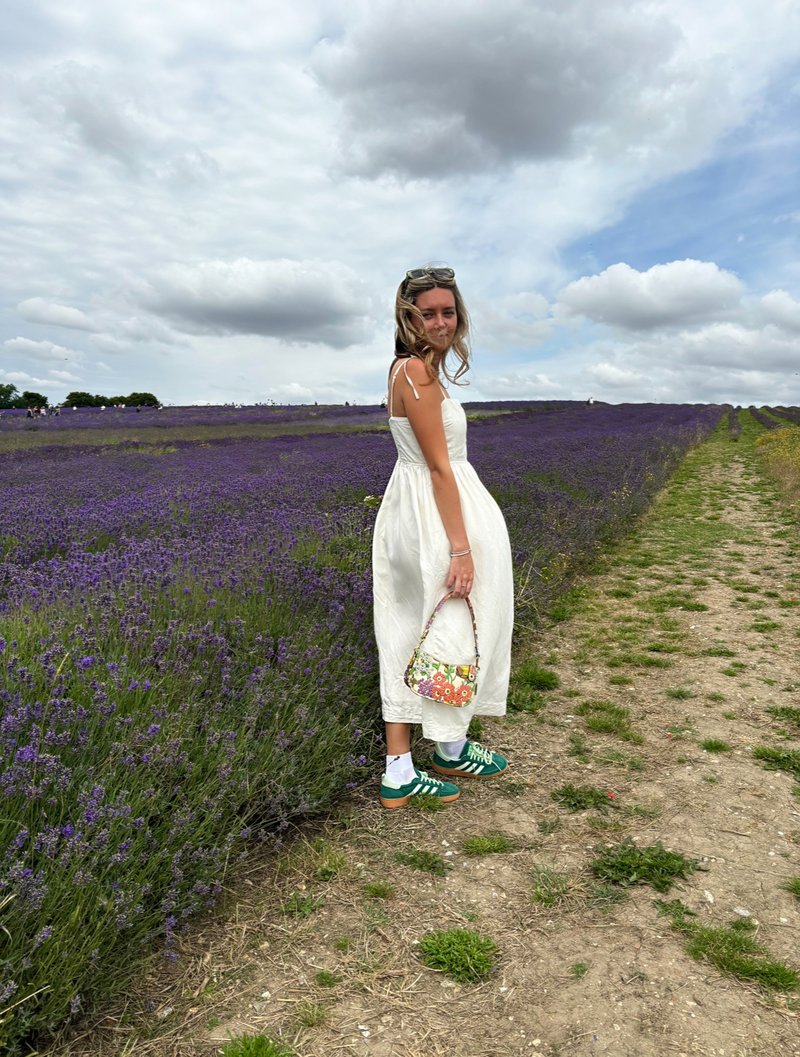 Photo of a young white woman in a white dress, in a field of purple lavender. She is looking over her shoulder at the camera, and holds a multi-coloured floral bag in her hand. Ahead of her are rows of lavender plants, and a blue and grey sky.