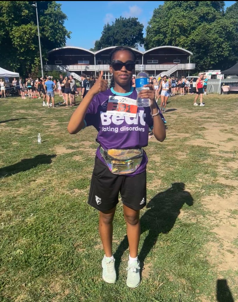 Photo of a young black woman in sportswear on a field. She wears sunglasses, a purple Beat-branded t-shirt and shorts. She holds a water bottle in one hand and has her thumbs up on the other.