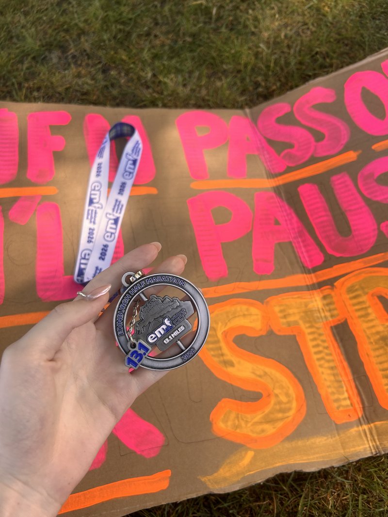 Hand holding a silver medal, which reads 'EMF' and 'Edinburgh Marathon Festival'. Behind the hand and medal is a handmade sign on cardboard.