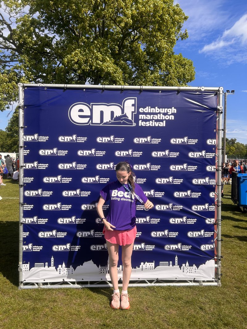 Young white woman stands in front of a banner that has the Edinburgh Marathon Festival logos across it. She wears a purple Beat-branded t-shirt, and holds up a medal from around her neck.