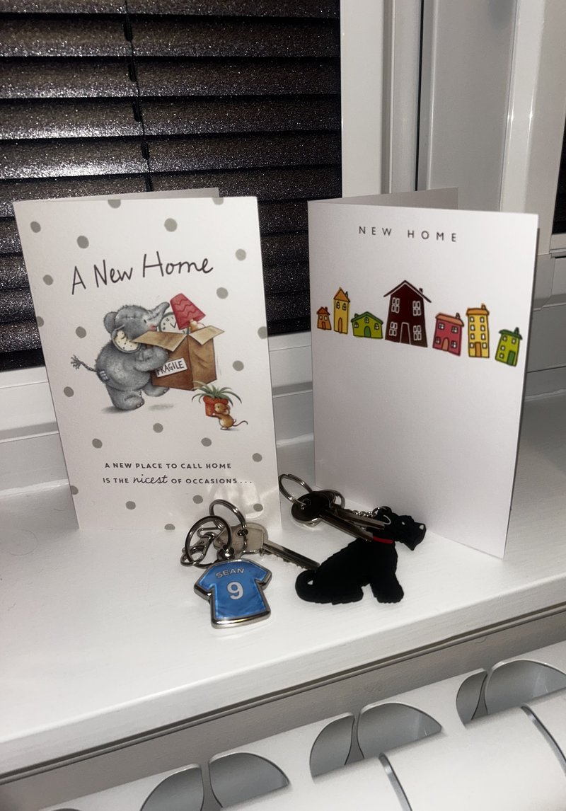 Two cards on a windowsill have 'New Home' messaging. In front of them are two sets of house keys.