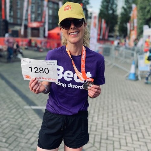 Woman smiling and holding her event number and a medal, wearing a purple Beat t-shirt and a yellow cap that says Summer Walk