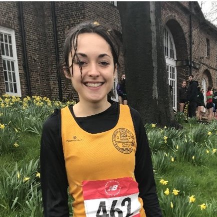A smiling picture of Lucy standing in front of a patch of grass and daffodils, with a brick building in the background. She is wearing a black t shirt under an orange vest with a race number attached to it.