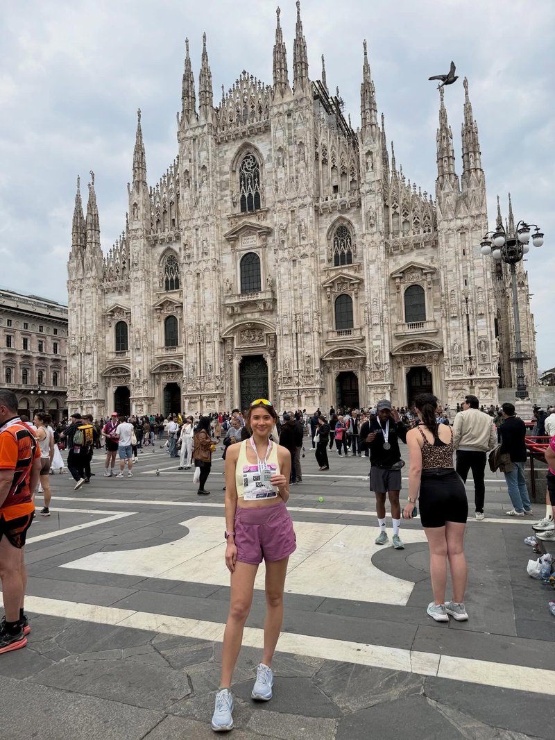 Young woman stands in front of a large, grand cathedral structure behind her. She wears running shorts and top, and is holding a medal around her neck. Surrounding her are people looking or taking photos of the catherdral.
