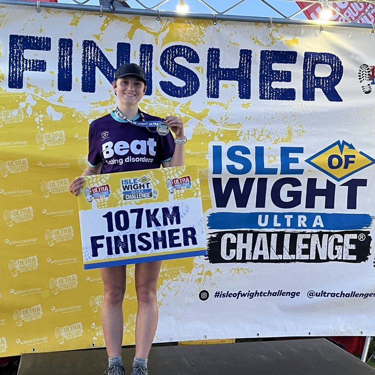 Individual wearing a Beat t-shirt whilst holding a medal and a sign saying '107km finisher'.