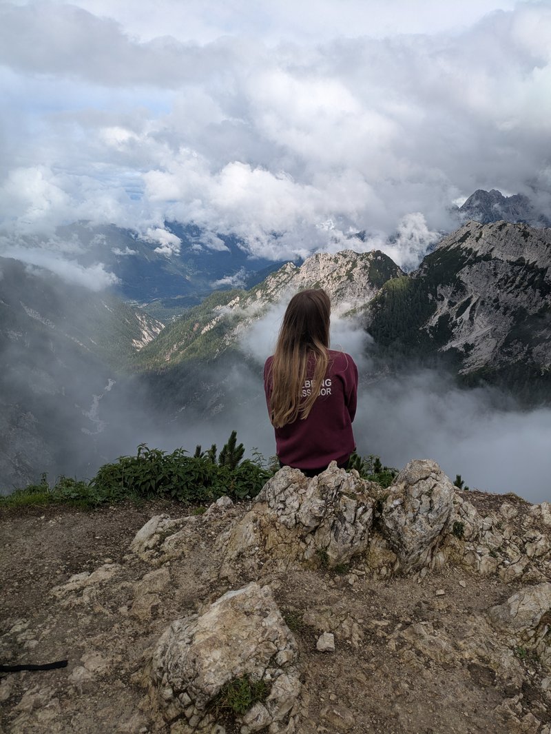 Young woman sitting on a cliff, looking out towards a beautiful and sublime mountainous landscape in front. Three mountain peaks are shrouded by clouds, and stretch out far into the landscape.