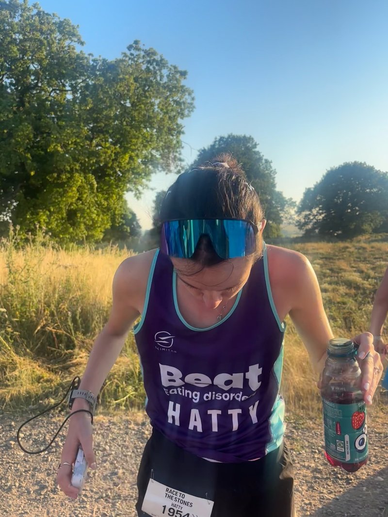 Young white woman looks down, showing the reflective sunglasses on her head. She wears a dark purple Beat branded purple t-shirt, with her name 'Hatty' across it. Behind her are fields of grass and trees.