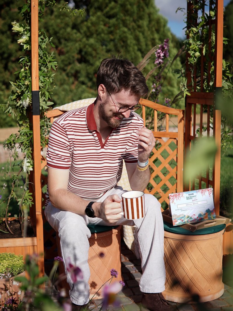 Young white man smiles and sits on a small wooden bench, holding a striped mug matching his striped shirt. Surrounding him is flowers and green plants.