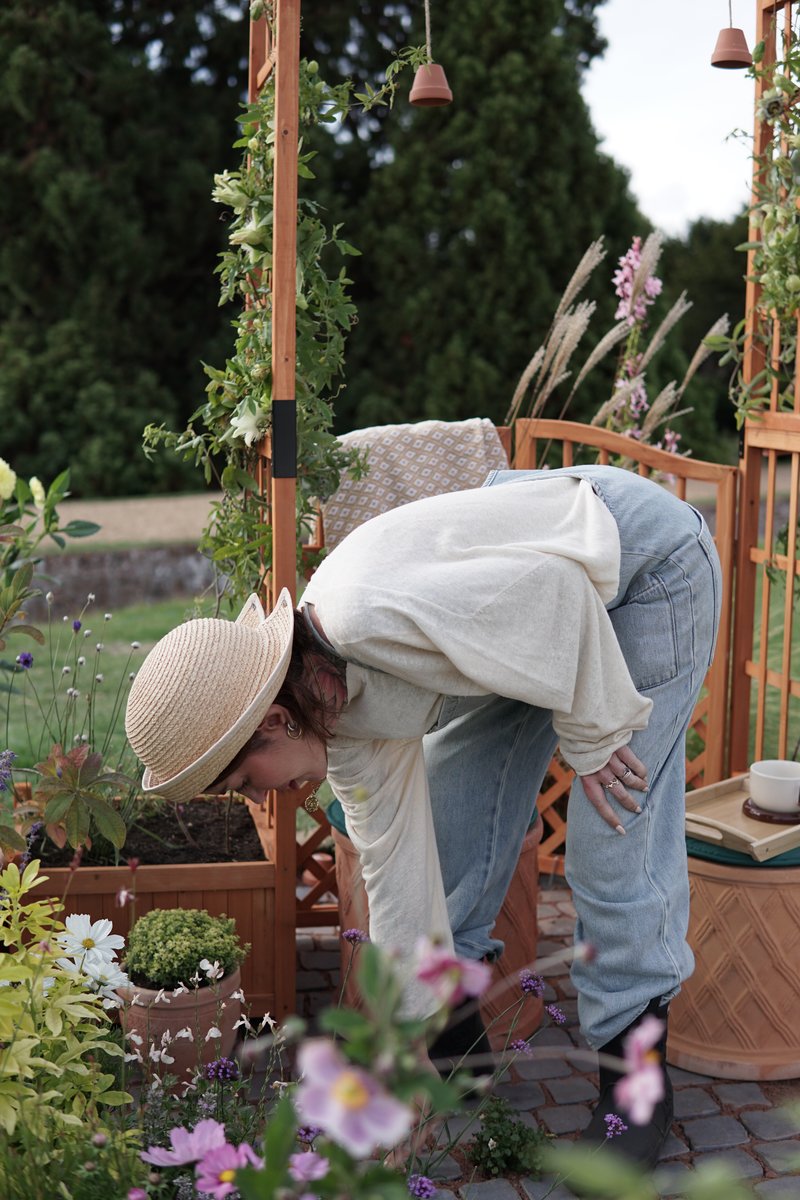Young white woman bends down, looking at some flowers in front of her. She wears a straw summer hat, long sleeves and jeans. Surrounding her are flower pots, a wooden bench and pergola.