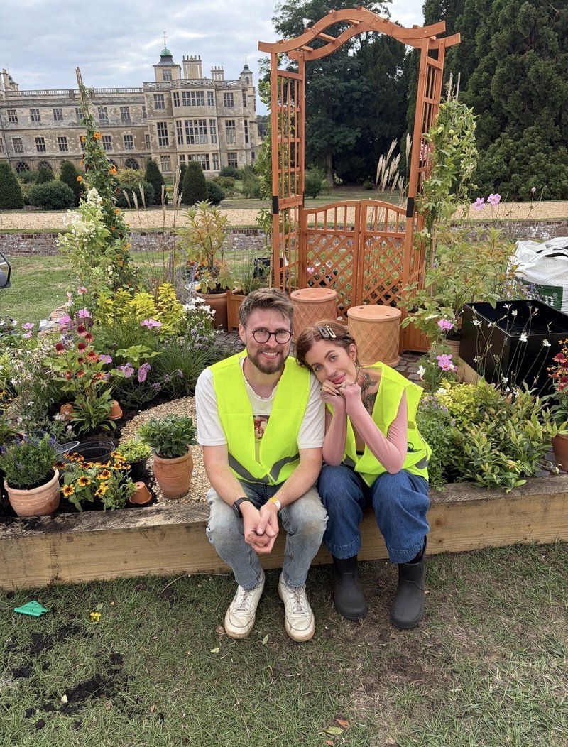 A young white couple sit together, in front of a garden area, filled with flowerpots and plants. Behind this is a grand old-looking house.