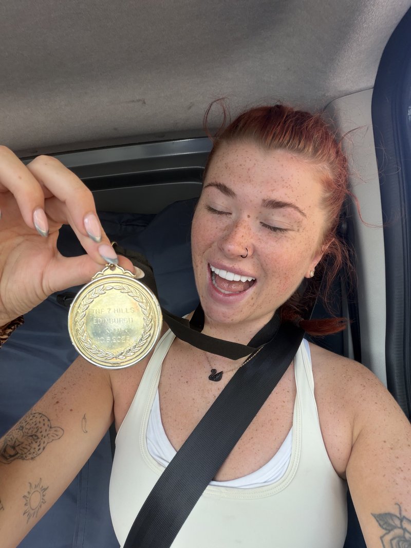 Young white woman with red hair and freckled skin smiles wide, holding up her gold medal from around her neck. She wears a sports top.