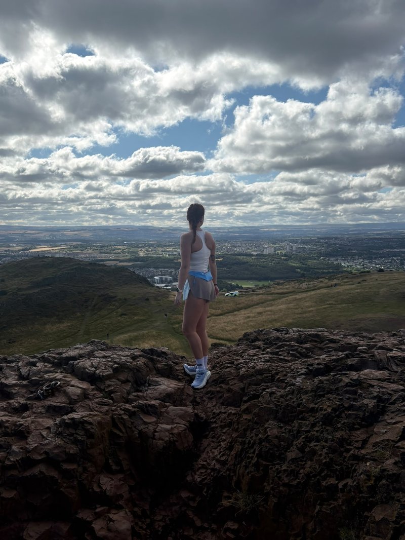 Young white woman in sportswear stands on a rock, overlooking green hills and an old-looking town below. Fluffy clouds fill the blue sky above.