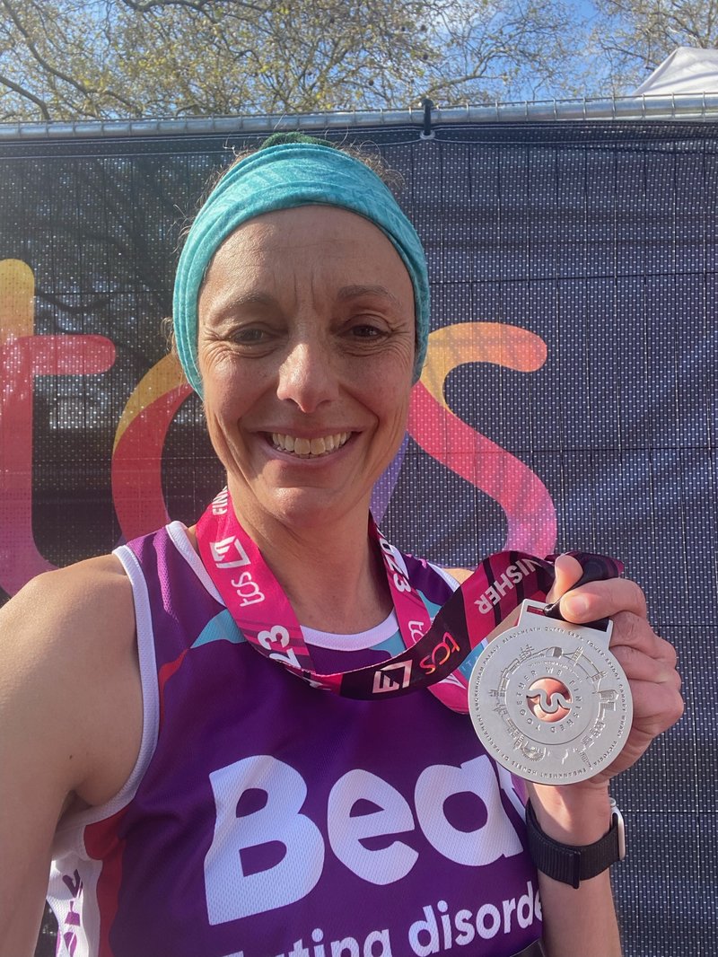 Selfie photo of a white woman smiling into the camera, holding up her London Marathon gold medal to the camera. She wears a teal headband and a purple Beat-branded vest.