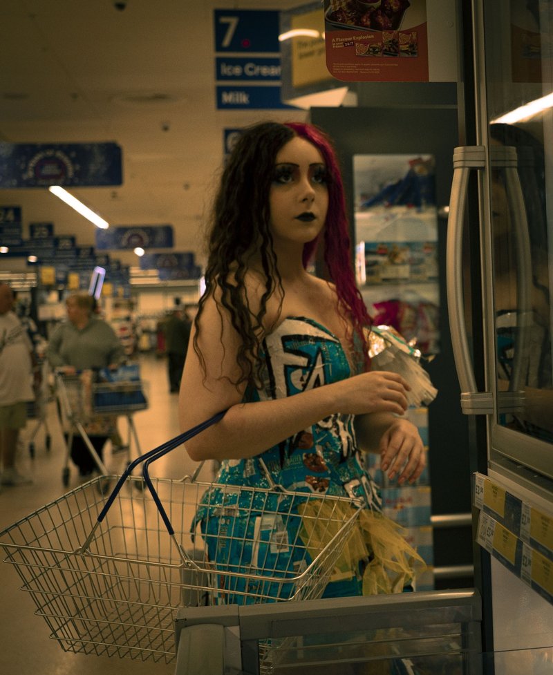 Photo of a young woman in the isle of a Tesco supermarket, near a freezer door that she is looking at. She has split black and red hair, with dark eye makeup and black lipstick. She is wearing a teel dress made of food wrappers and tulle. She holds a supermarket basket in the crook of her arm.