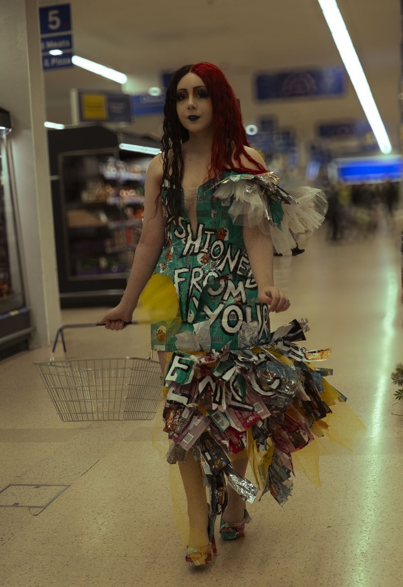 Photo of a young woman walking through the middle isle of a Tesco supermarket. She has split black and red hair, and matching red and black makeup. She wears a teal dress made of wrappers, that goes down into a skirt of coloured wrappers. It reads &#x27;fashioned from your expectations&#x27; across the body of the dress. She also wears tall high heels and holds a shopping basket in one hand.