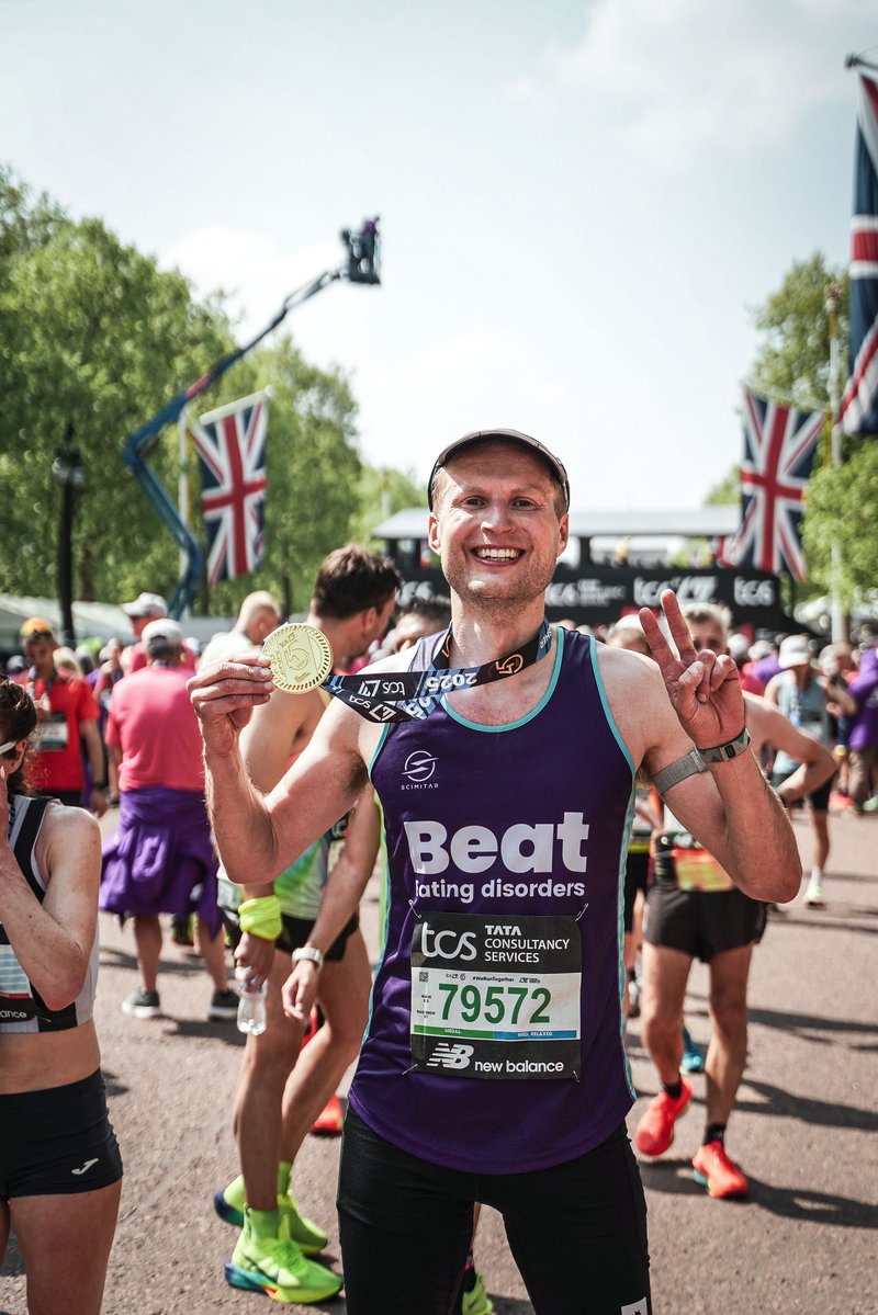 Young white man smiling at the camera, holding up his gold medal around his neck in one hand, and a peace sign with his other. He wears a cap, and a dark purple Beat branded vest. Behind him is a crowd of other runners, and union jack flags lining the pavement.