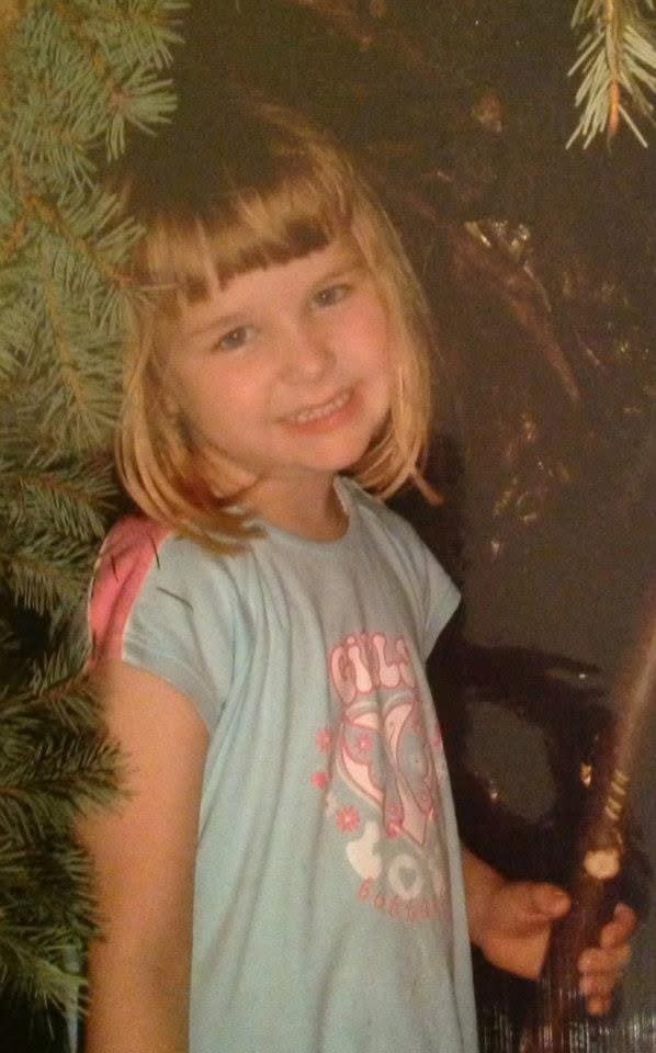 Photo taken from a disposable camera of a young white girl smiling at the camera, under a fir tree. Her hair is in a blonde bob and she has light eyes and a baby-teeth smile. Her t-shirt is light blue and pink.