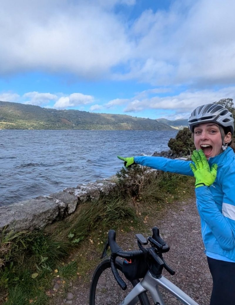 Photo of a young white woman looking at the camera, with one hand over her open mouth, and one hand pointing out towards the lake in front of her. She wears a silver bike helmet, blue cycling jacket and neon gloves. Across the water is forested land, and above is a cloudy blue sky.