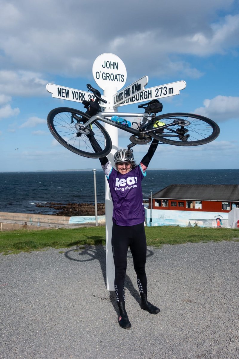 Photo of a young white woman smiling, holding up a bike over her head. She wears a silver helmet, cycling gear and a dark purple Beat-branded t-shirt. She stands in front of a old looking white sign that reads &#x27;John O&#x27;Groats&#x27;. The sign has other branches pointing towards New York, Edinburgh and Lands End. Behind the woman is the tea and a cloudy blue sky.