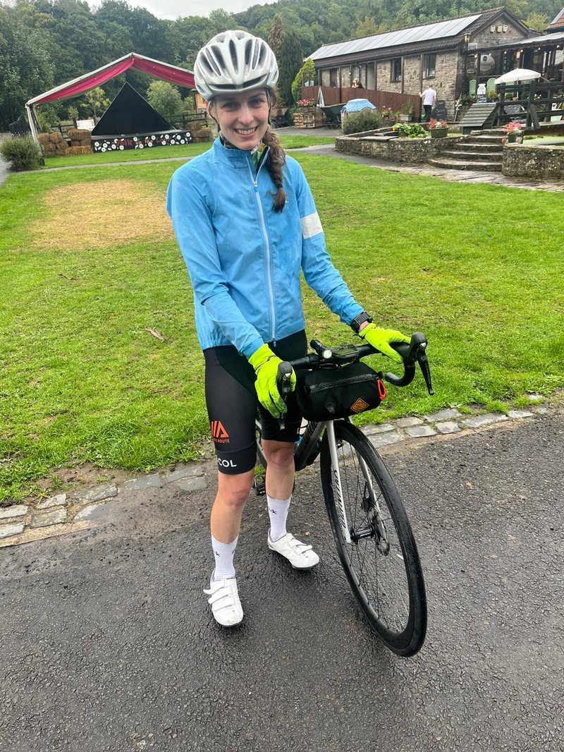 Photo of a young white woman smiling at the camera, and standing with her bike. She wears a silver helmet, a light blue light-weight jacket, neon yellow gloves, and black cycling shorts. Behind her is a lawn, with a forest further back.