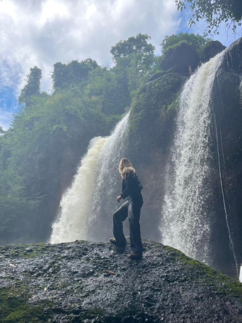 Photo of a young woman with blonde hair, looking over her shoulder at two waterfalls behind her. She stands on a wet rock, wearing black jeans and a black jacket. Behind her, the scenery is lush and green.