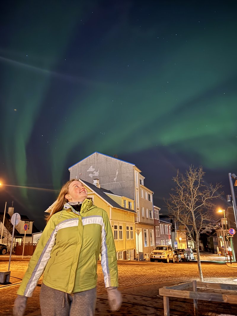 Young white woman in a green reflective coat, looks up at a green streaks of the Northern Lights in a dark sky. Behind her is a street with snow on the roads.