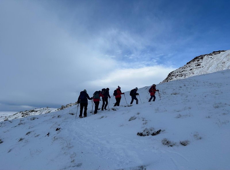Six people walk in a line across the snow. They are all wearing puffy coats, trousers, hats and walking sticks. Surrounding them is snow, mountains and wispy clouds.