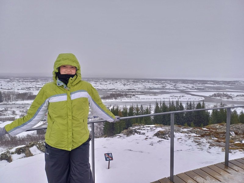 Young white woman in a thick green coat, smiles, standing in front of a flat snowy landscape. Barren roads stretch out into a grey sky, and tall fir trees line an area on the right hand side.