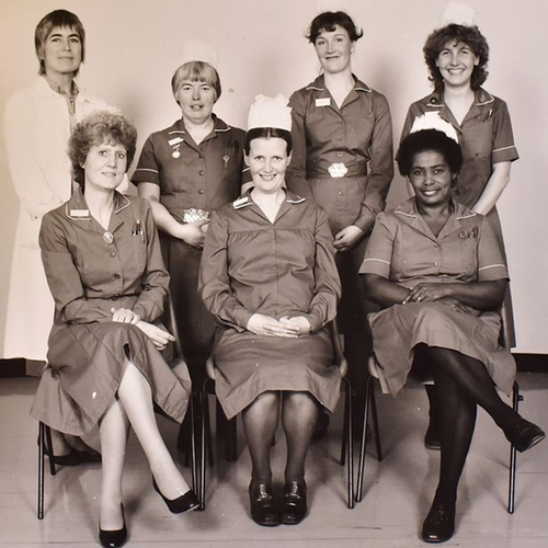 A black and white photo of a group of nurses sitting posing for the photo. May Tanner is sat on the far right, smiling at the camera with her legs crossed. She is the only Black nurse; the rest of the women in the photo appear to be white.