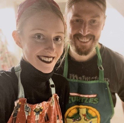 A smiling selfie of Yan and Ross. They are both wearing black t shirts, with patterned aprons.