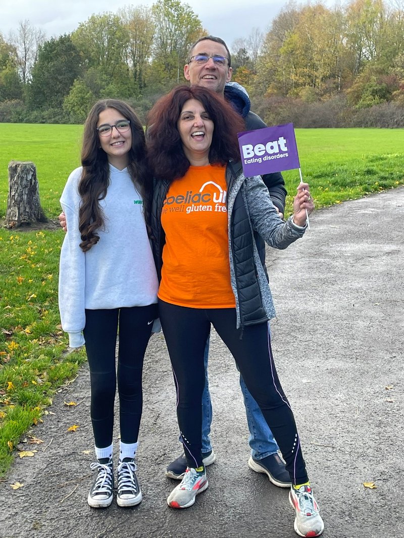 Photo of a family of three smiling at the camera, in a park. A daughter on the left is next to her mum on the right, with the father behind them. The daughter has long brown curled hair, glasses, and a light grey hoodie on. The mother has dark red-dyed hair, and wears an orange Coeliac UK branded t-shirt, under an open coat. In her hand is a purple paper flag with the Beat logo on it. The father has brown short hair and wears glasses.