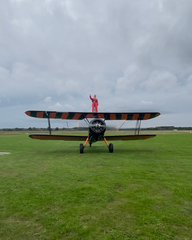 Person stands on top of a biplane, with it's propeller spinning. They have their hand up waving, and are in an orange jumpsuit. The plane is in a grassy field, and the sky is cloudy.