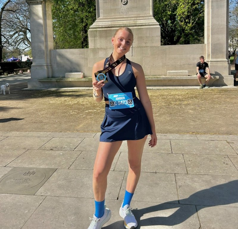 Photo of a young white smiling woman, holding up a medal from around her neck. She is outdoors in the sunshine, with a statue behind her. She has blonde hair and wears a navy sports top and skirt.