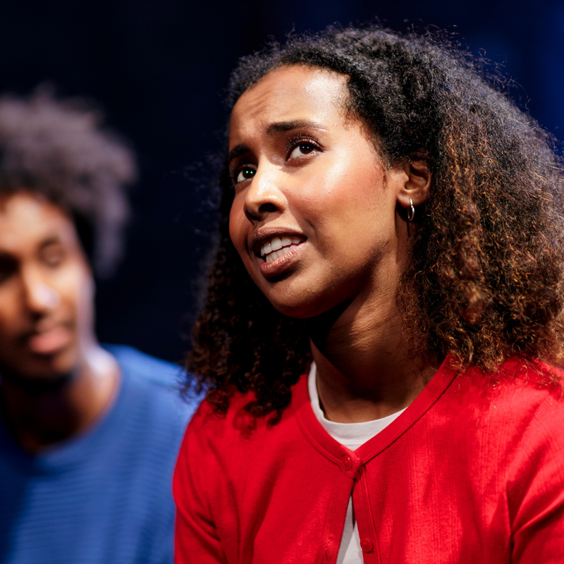 Young black woman looks off to the right corner, mid-speech. She has shoulder-length black coily hair, and wears a red cardigan. In the background, with a blur, is a black man looking at her.