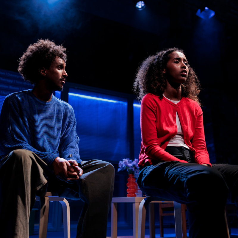 Two young black people sit in chairs on a stage, acting. The man on the left looks concerned at the woman on the right. He wears a blue jumper, and has his hands clasped in his lap. The woman on the right has shoulder-length coily black hair, and wears a red cardigan.